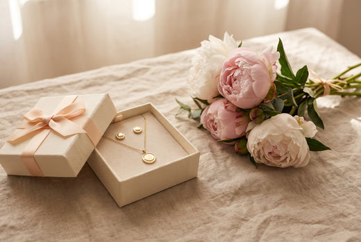 Gold-plated necklace and earrings in an open gift box beside pink peonies on linen