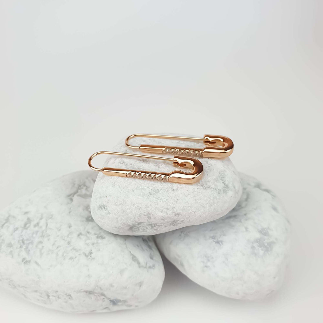 Pair of rose gold-tone safety pin earrings on white pebbles, clasp visible