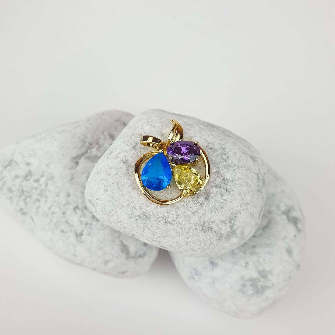Gold-tone apple pendant with three bright stones at an angle on pale pebbles