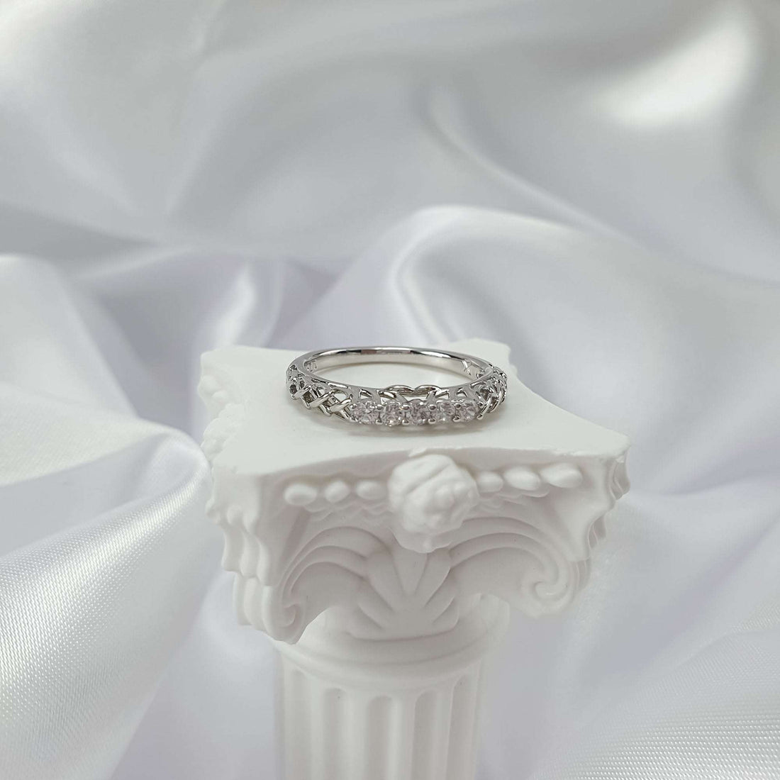 Silver-tone openwork ring on a white column pedestal with a satin backdrop