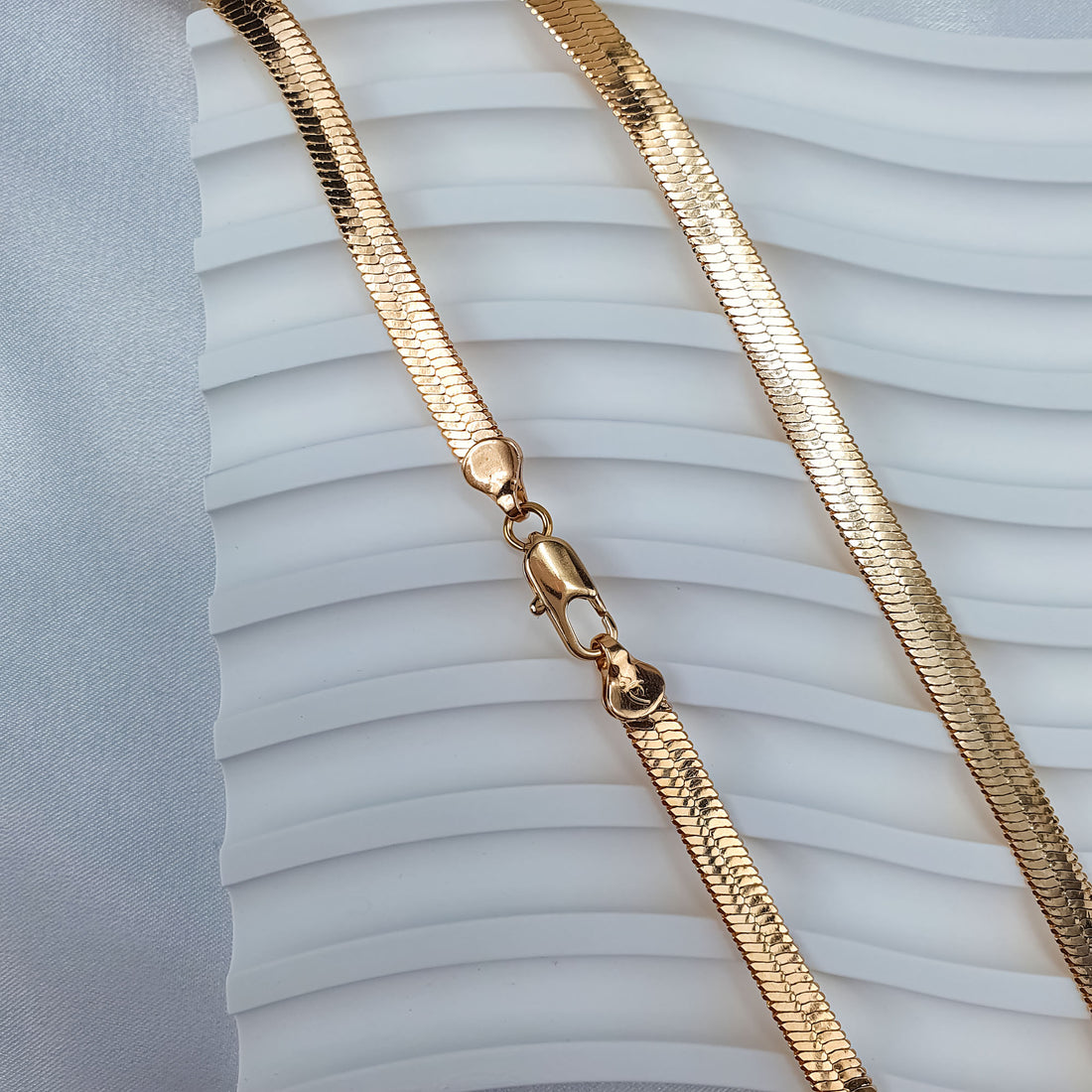 Close-up of a flat gold-plated chain on a wavy white stand, lobster clasp visible