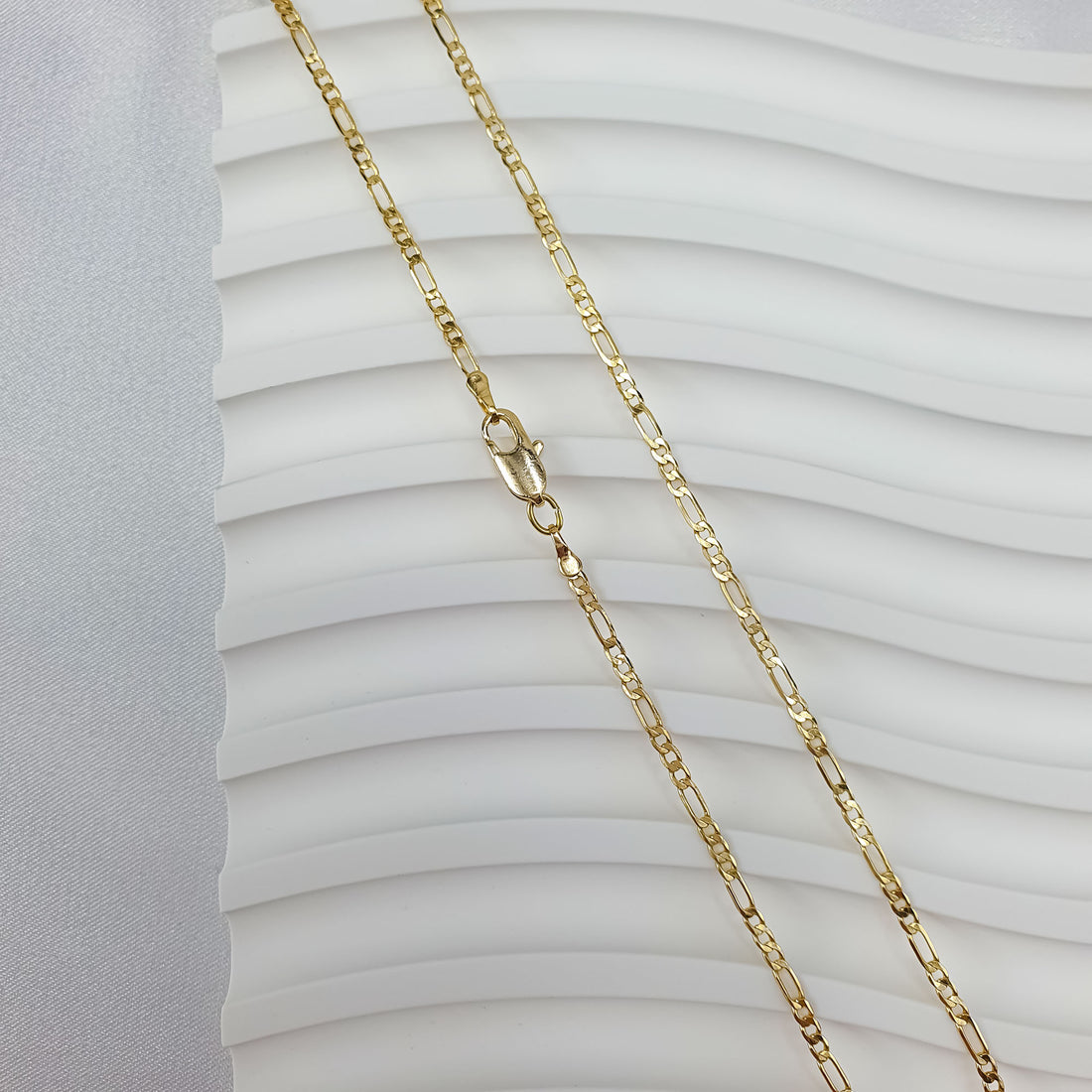 Fine gold-tone Figaro chain laid out on a white wavy background