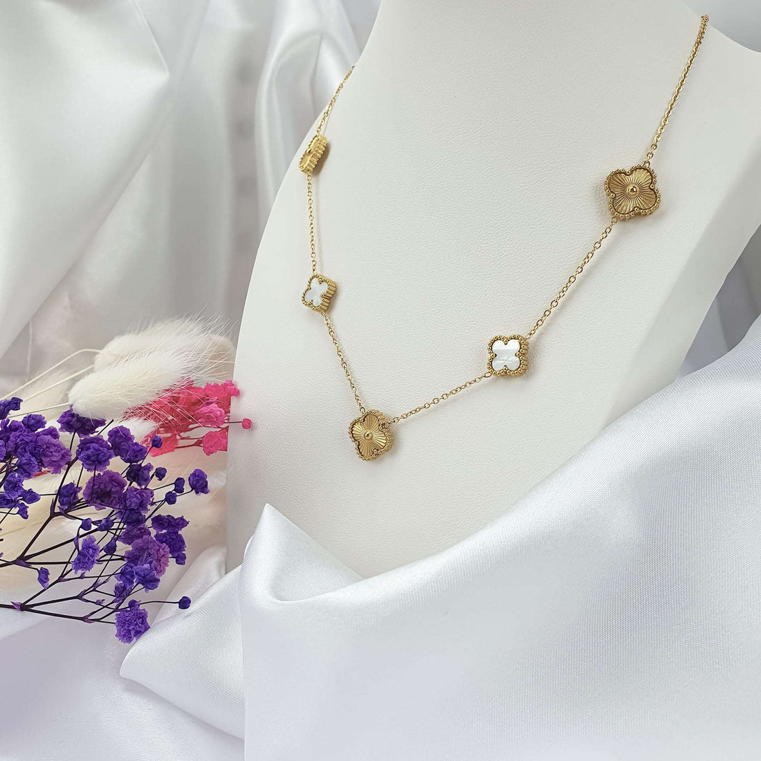 Gold-plated clover necklace on a white stand among satin and dried flowers