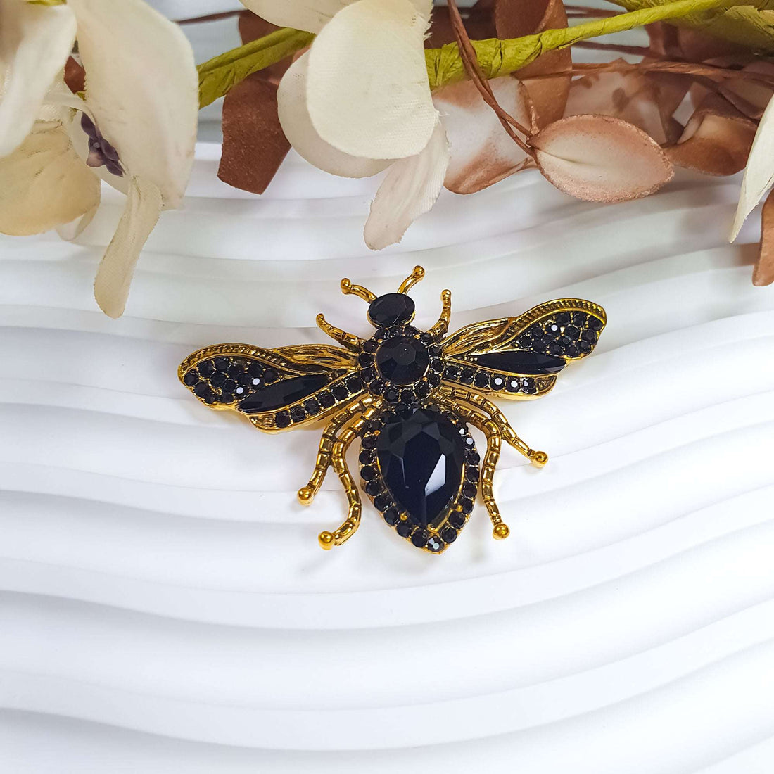 Gold-tone bee brooch with a black teardrop crystal on a wavy background