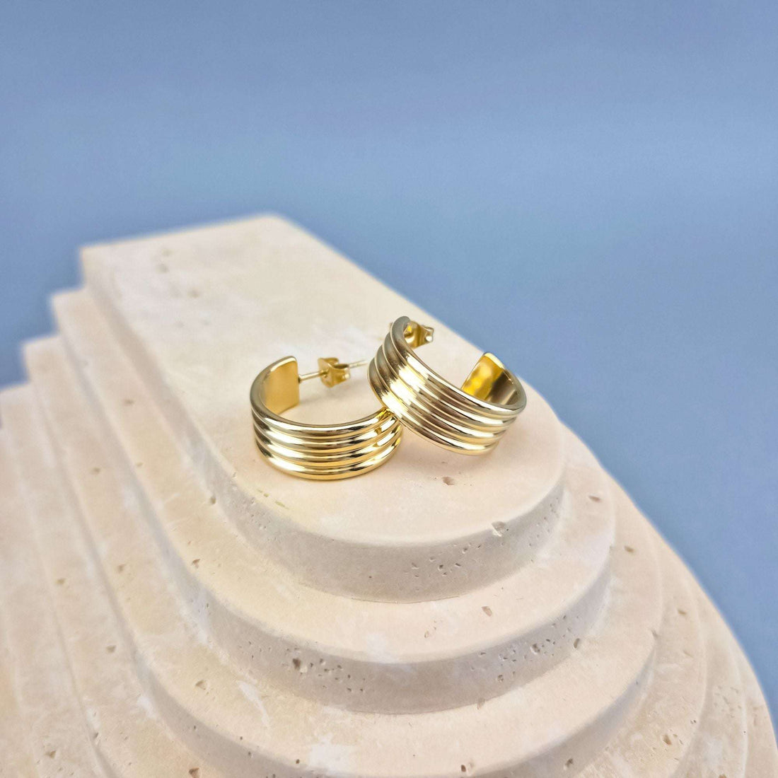 Gold-plated ribbed half-hoop earrings with push-back posts on a pale stone stand
