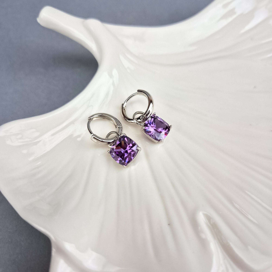 Silver-tone hoop earrings with sparkling violet square drops on a white ceramic leaf