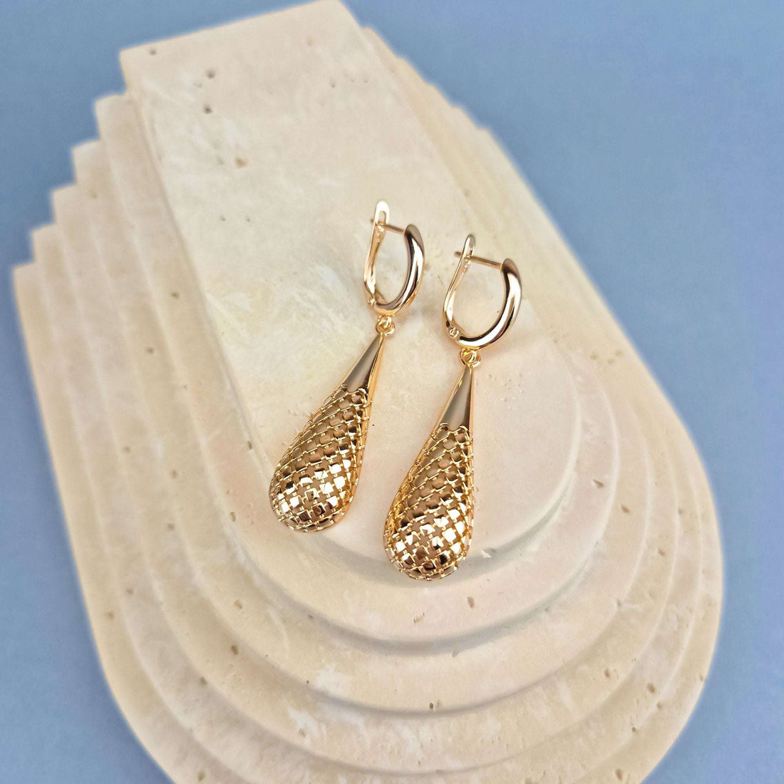 Gold-plated teardrop earrings on a pale stone stand against a blue background, leverback visible