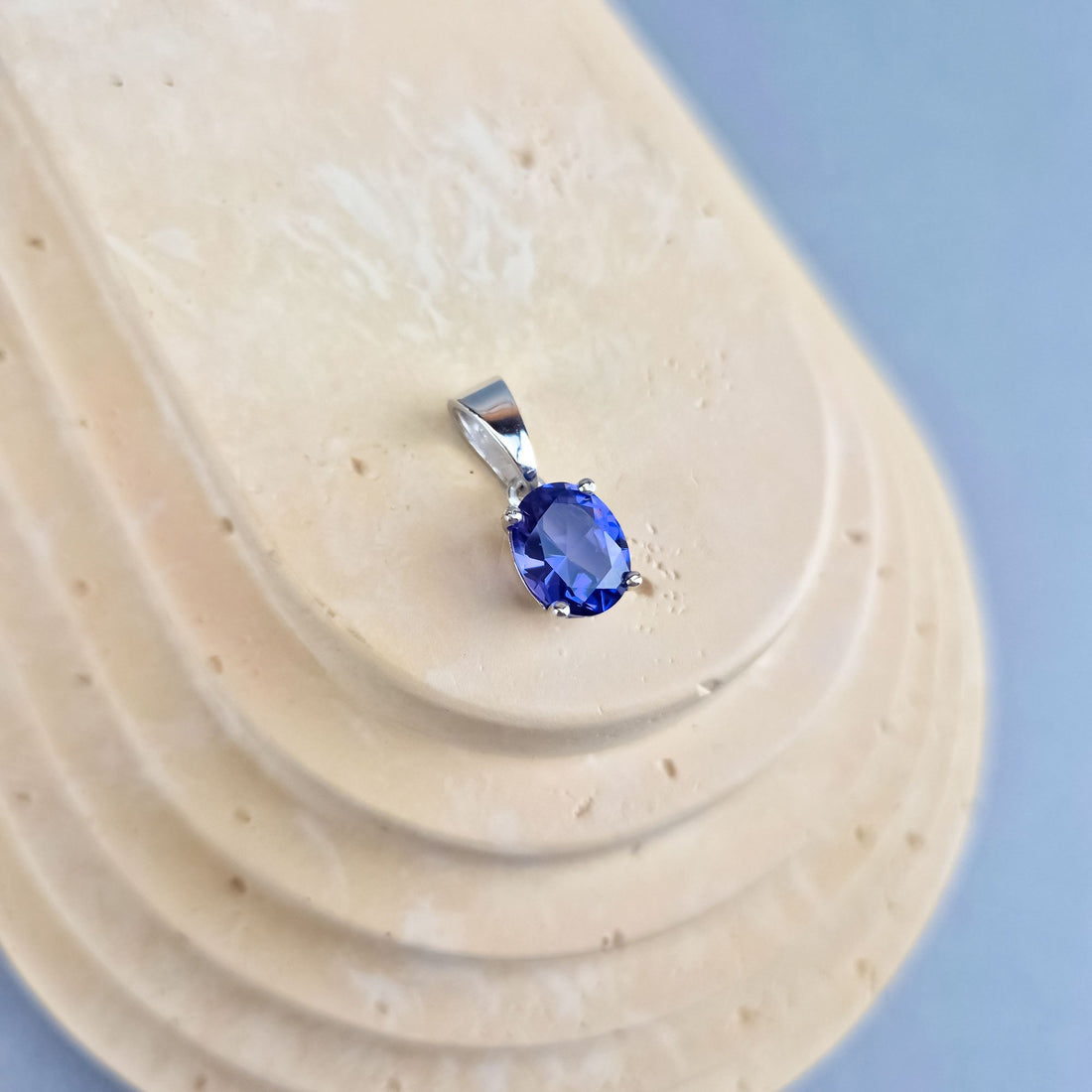 Blue-violet tanzanite pendant in silver tone on a beige stand, blue background