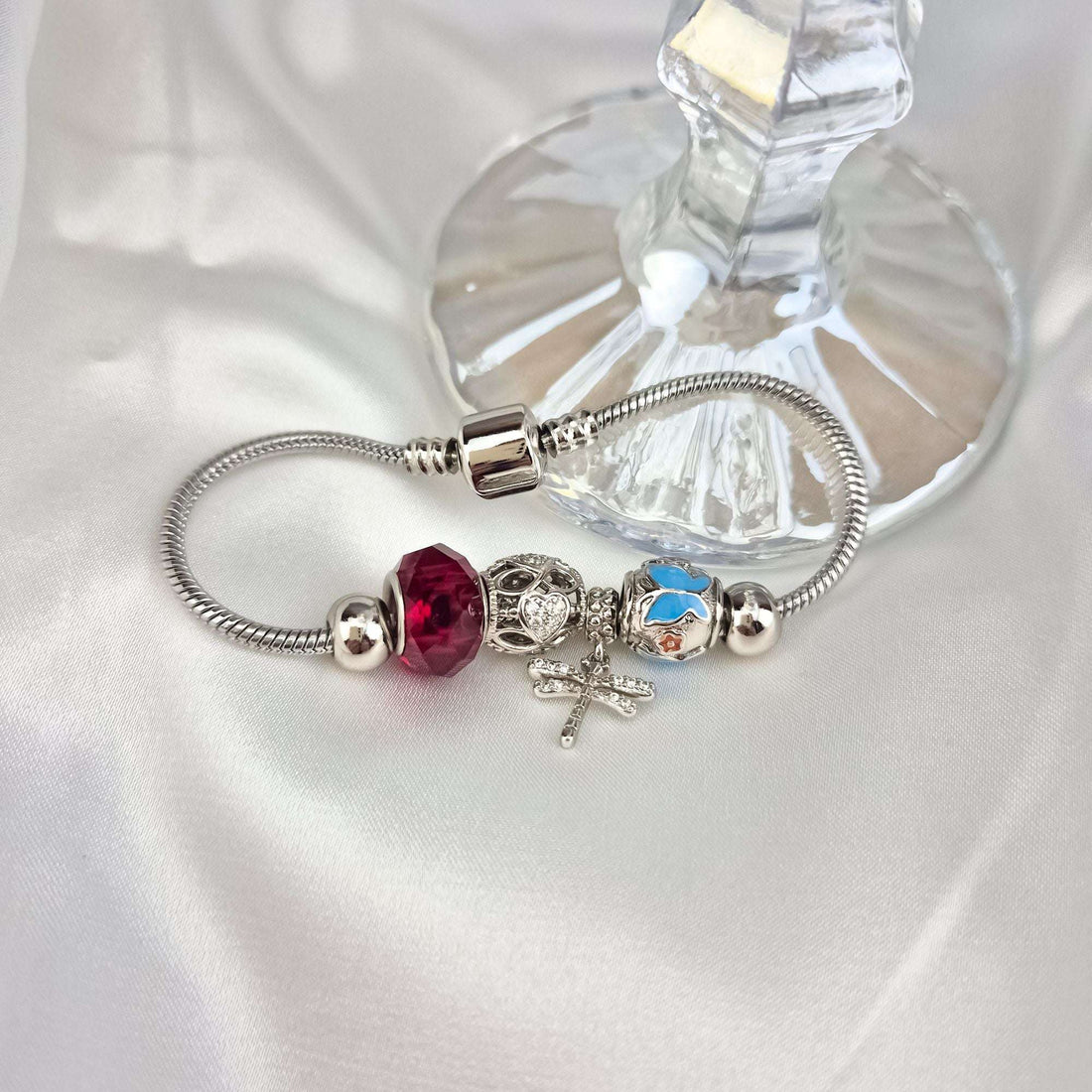 Silver-tone charm bracelet on white silk beside a crystal dish, snake chain visible