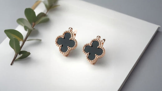 Rose gold-tone four-leaf clover earrings with black inlay on white paper beside greenery