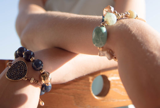 Crossed hands wearing stone bead bracelets, one with a black heart charm, in sunlight