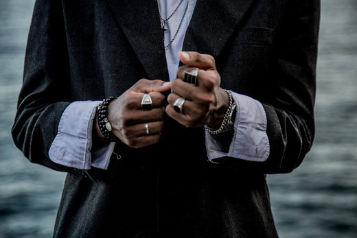 Man in dark jacket, hands clasped, wearing silver-tone signet rings and a chain bracelet