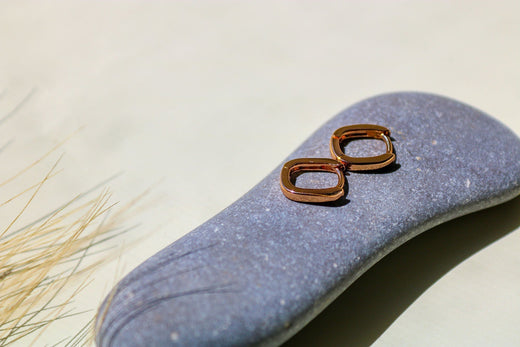 Two rose-gold-tone hoop earrings on a smooth sea pebble in sunlight