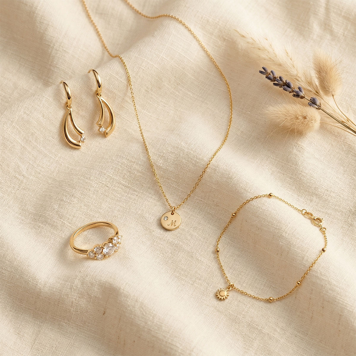 Gold-plated jewellery on cream linen: drop earrings, monogram pendant, stone-set ring and bracelet