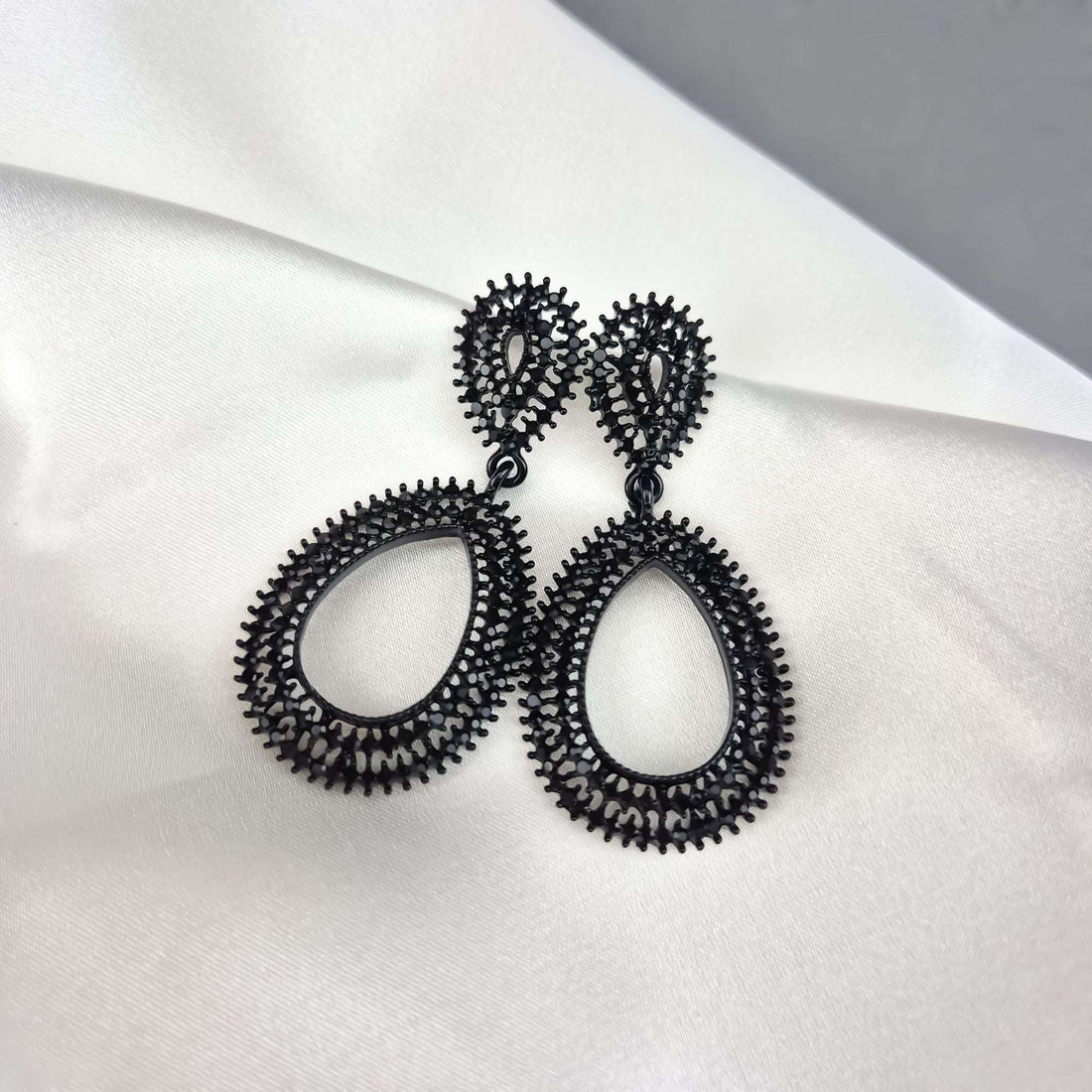 Two-part black openwork teardrop earrings at an angle on white silk drapery