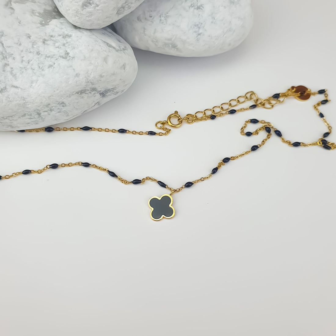 Gold-tone necklace with a black clover pendant laid beside stones, clasp visible