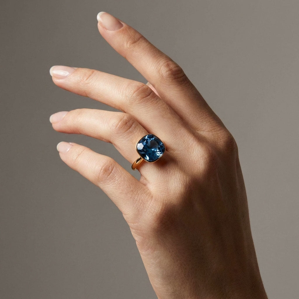 Model's hand wearing a gold-plated ring with a blue crystal, brown background