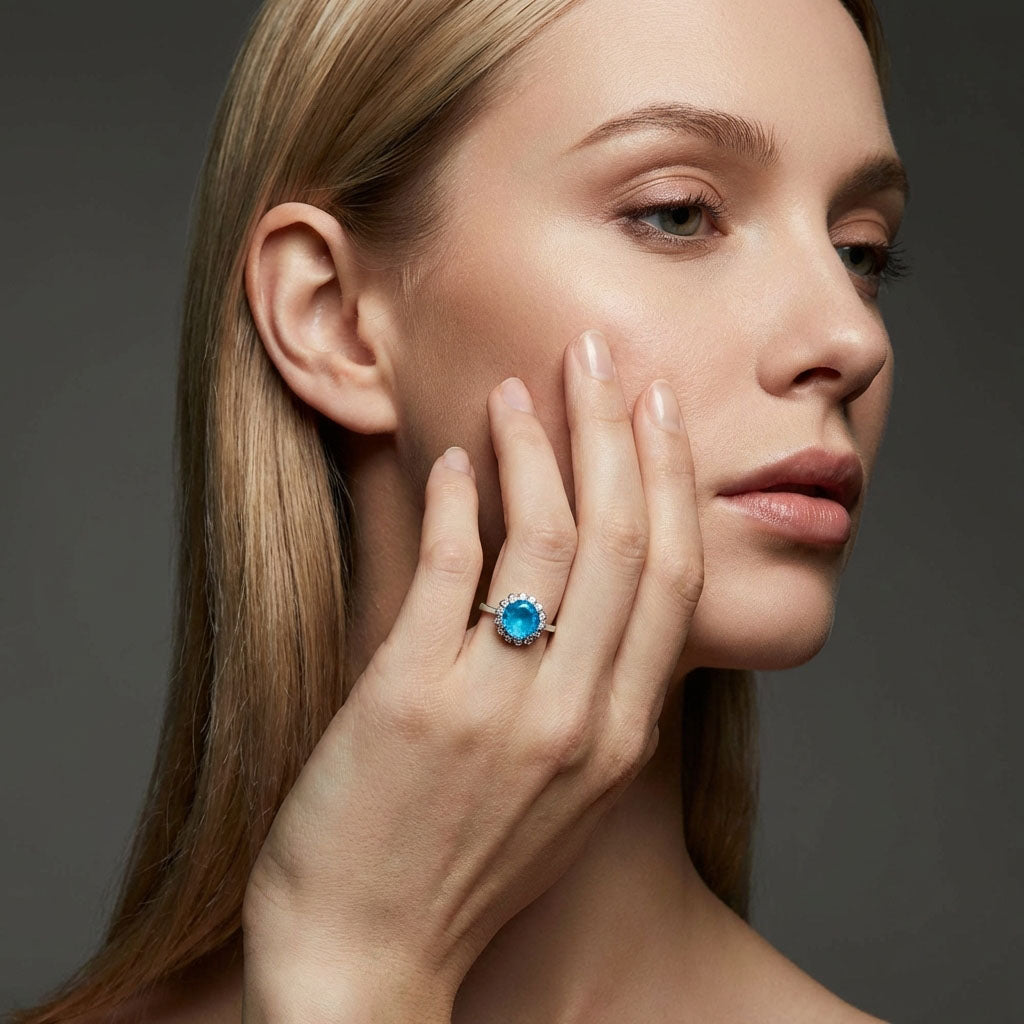 Model touching her cheek, wearing a silver-tone ring with a bright blue stone