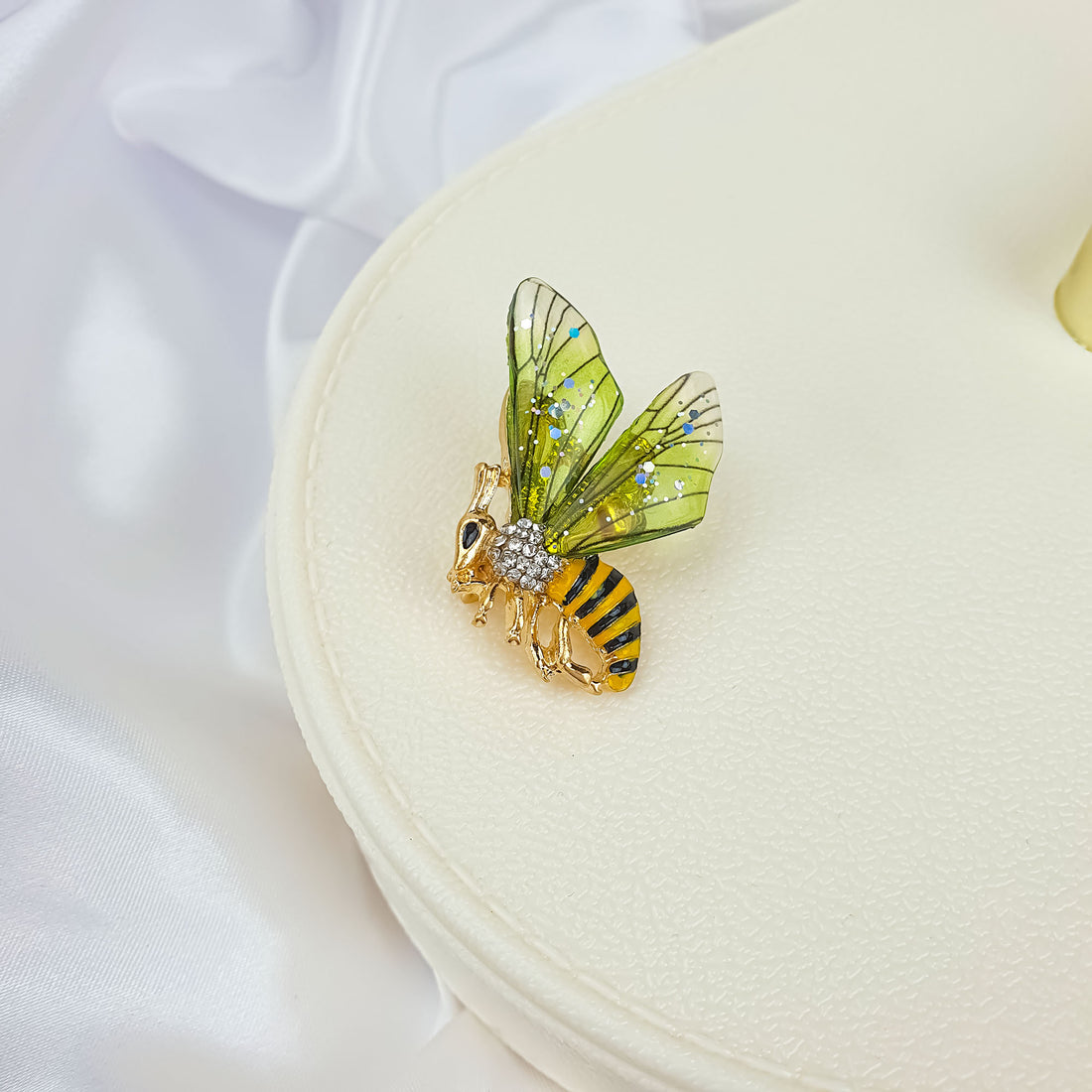 Gold-plated bee brooch on a cream jewellery box, green wings in close-up