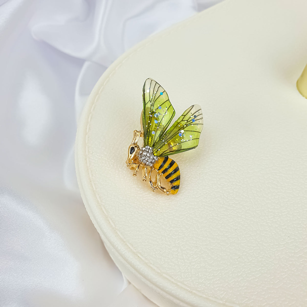 Gold-plated bee brooch on a cream jewellery box, green wings in close-up