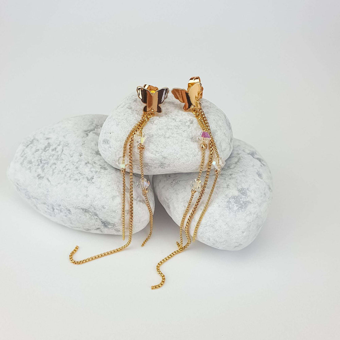 Gold-plated butterfly earrings with clear crystals and long chains on white pebbles
