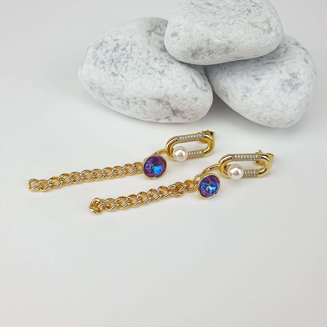Earrings laid out flat, gold-plated chains with violet-blue crystals, on a pale surface