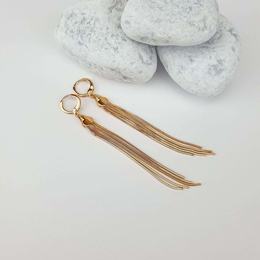 Gold-plated fringe earrings laid out beside white pebbles, their full length visible