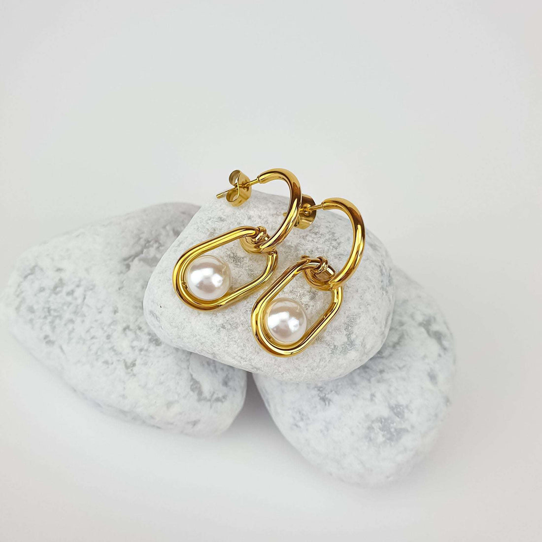 Curved gold-plated earrings with a faux pearl in an oval link, on white pebbles