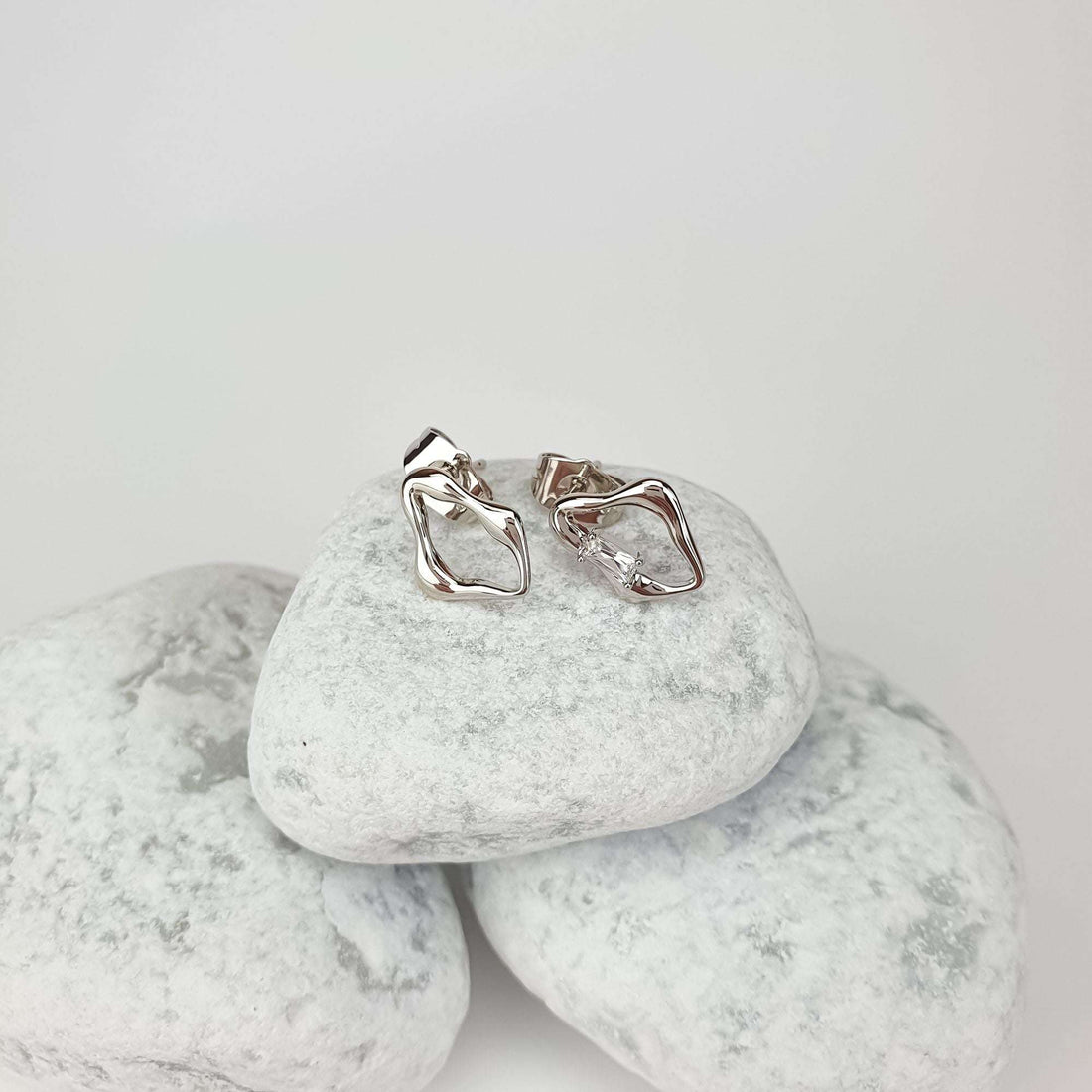 Asymmetric silver-tone earrings on a light stone, showing the glossy metal profile and crystal