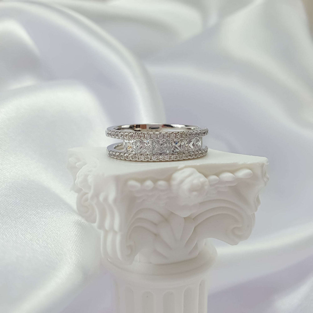 Silver-tone cubic zirconia ring from the side on a white plaster column, silk folds