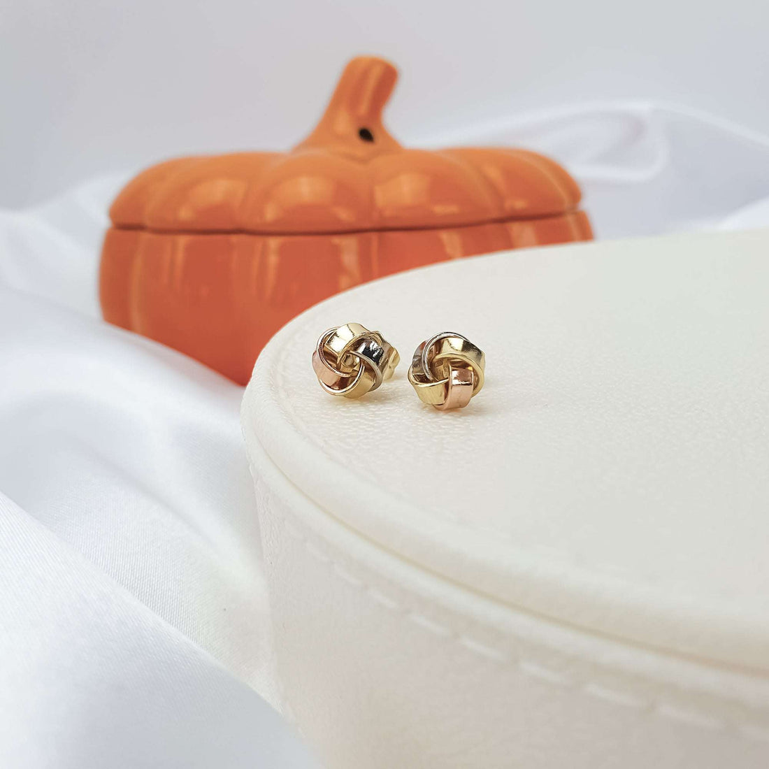 Tricolour knot studs in side view on a light jewellery box, ceramic pumpkin behind