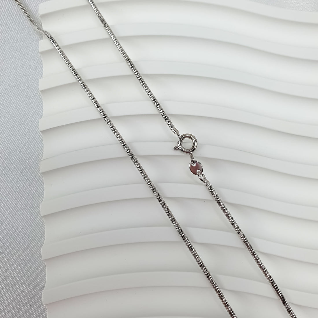 Smooth silver-tone snake chain laid on a white wavy surface, close up
