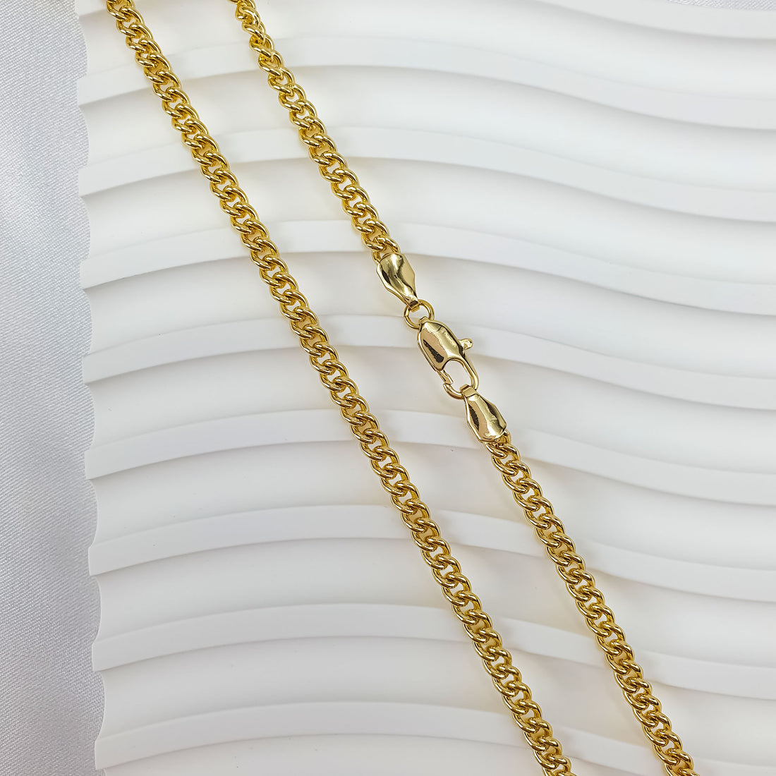 Polished gold-tone curb chain on a white wavy background with the clasp visible