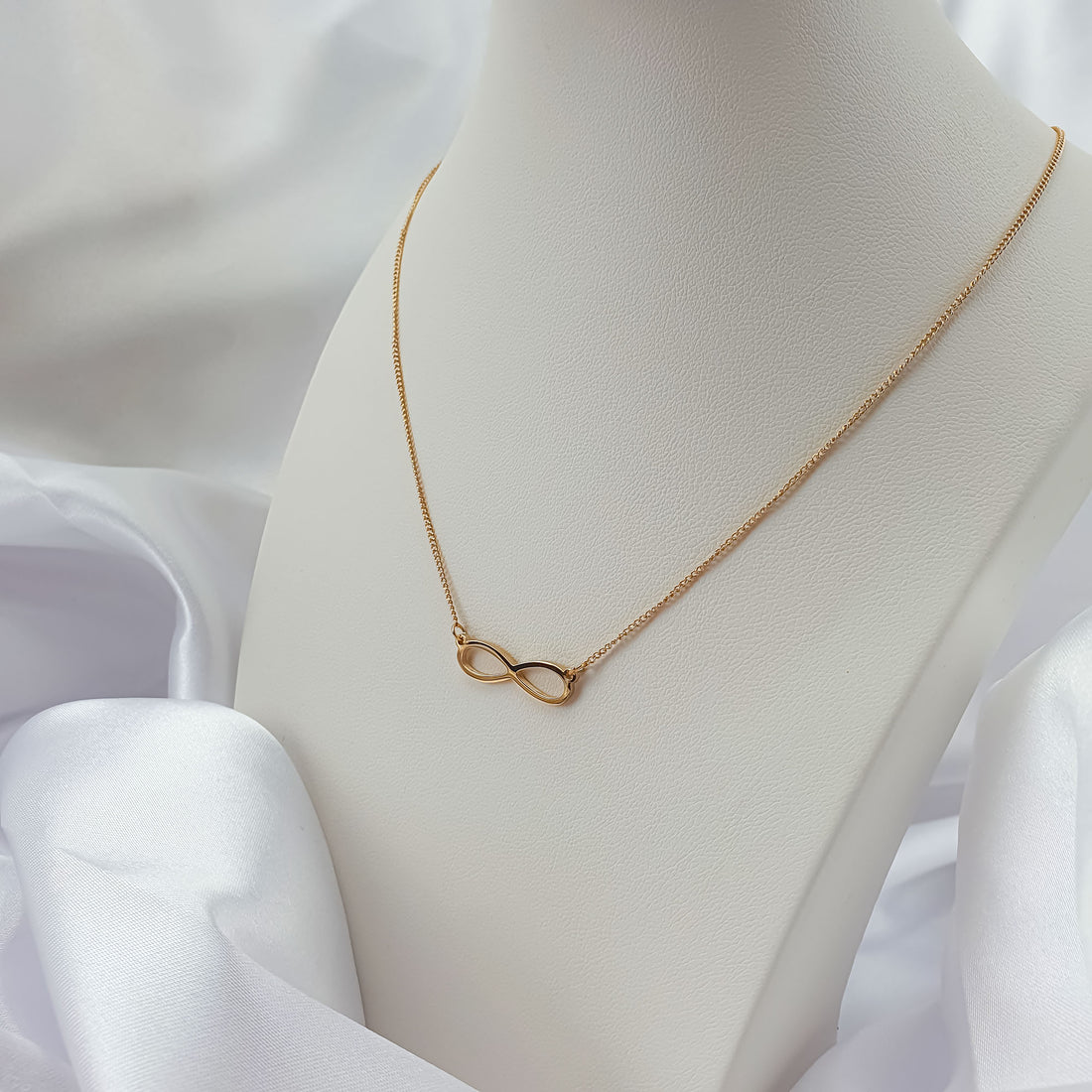 Gold-plated infinity necklace on a white bust with soft satin drapery