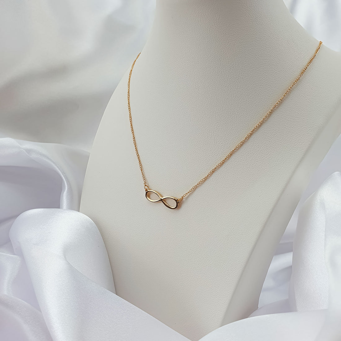 Delicate gold-plated infinity necklace on a pale bust, white satin behind