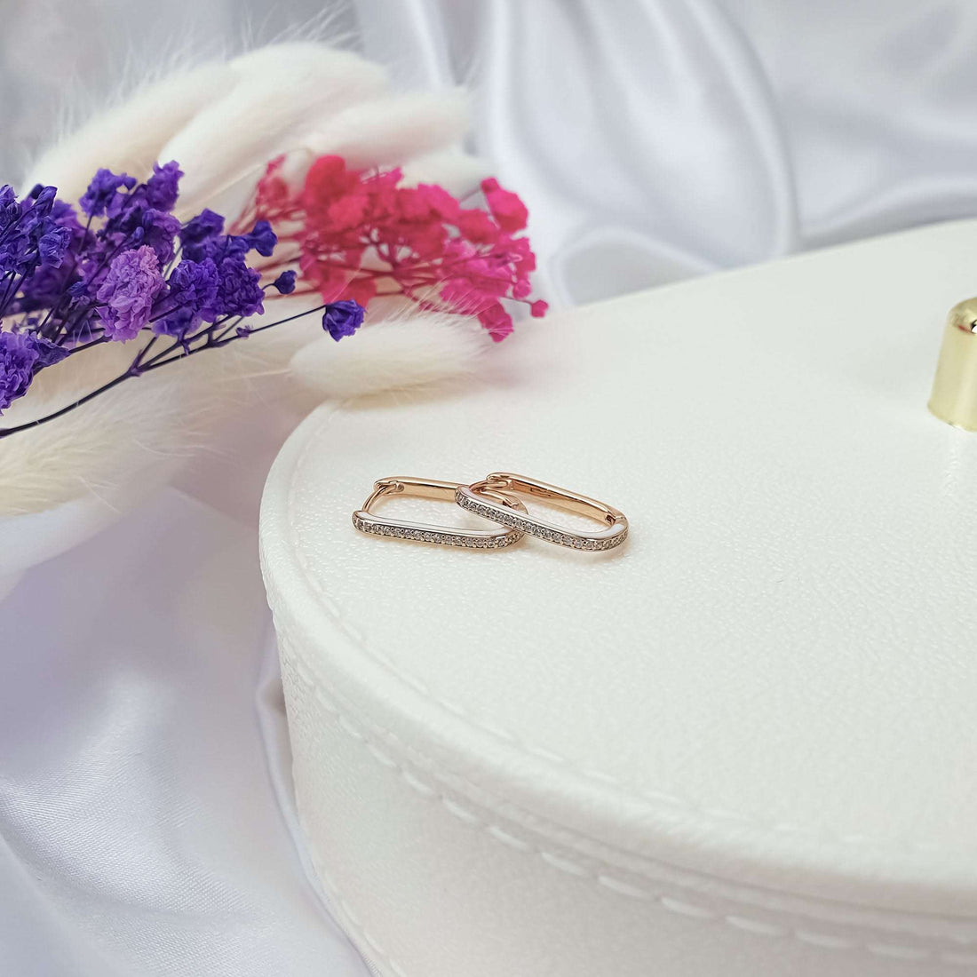 Rose gold-tone hoop earrings with cubic zirconia on a white box beside dried flowers