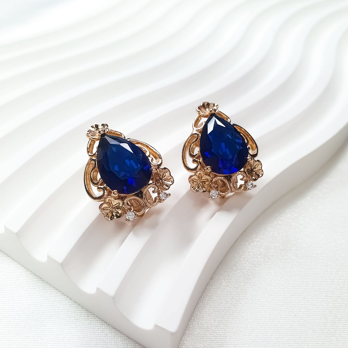 Dark blue teardrop clip-ons in gold tone on a white wavy stand, angled view