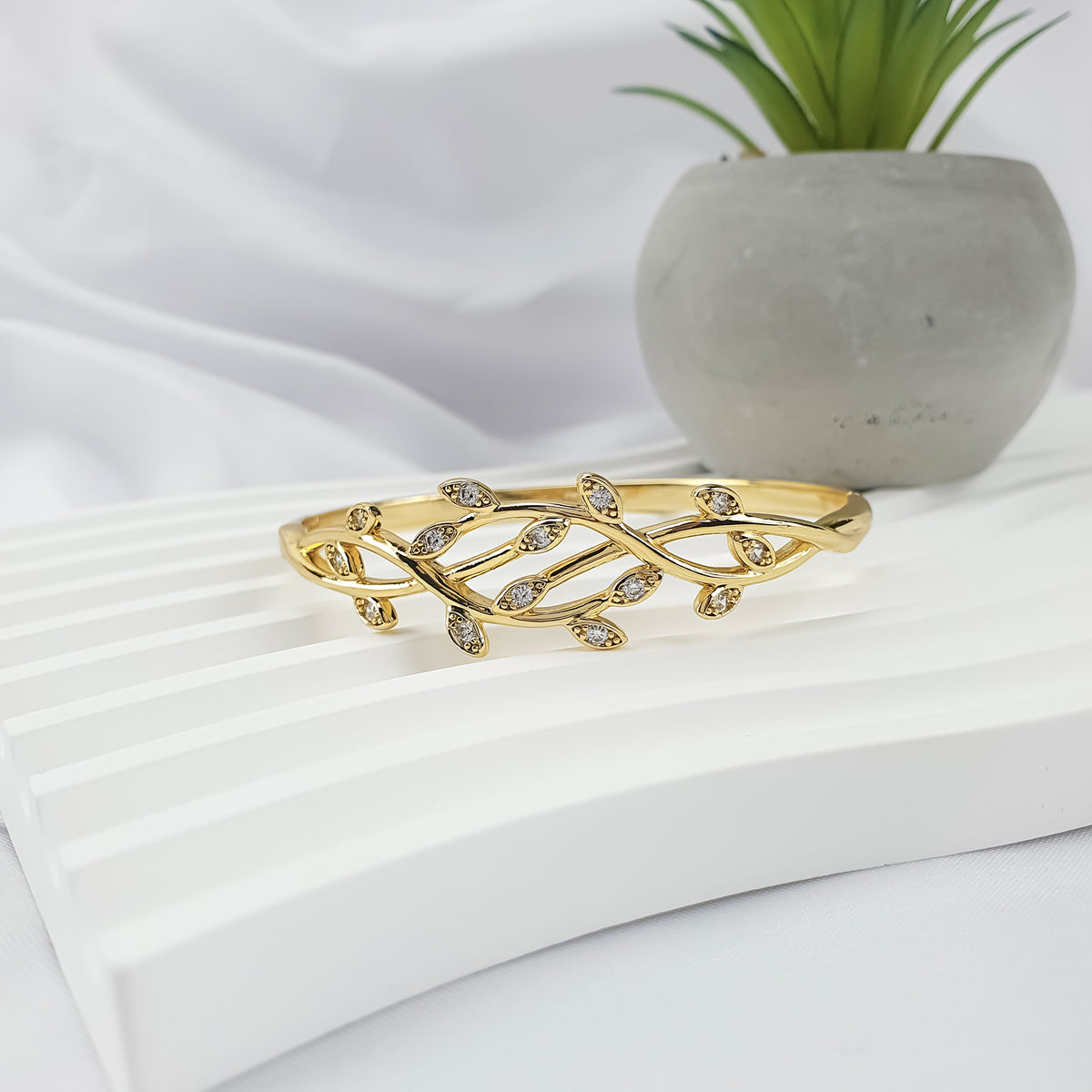 Gold-tone leaf bangle at an angle on a white stand beside a concrete pot