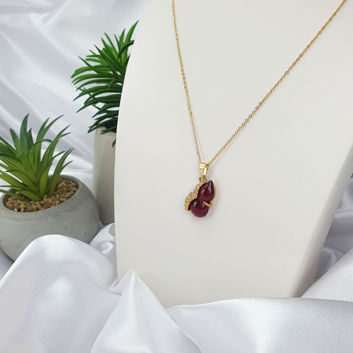 Gold-plated necklace with a dark red pendant on a white stand beside a plant