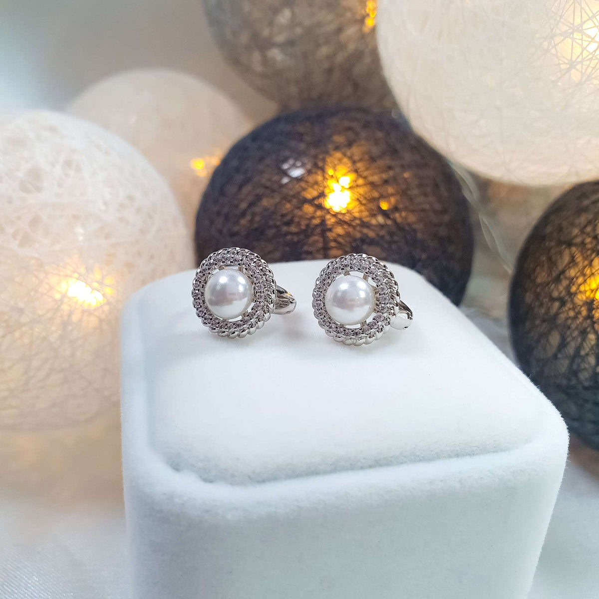 Silver-tone pearl earrings on a white velvet box, warm glowing lights behind