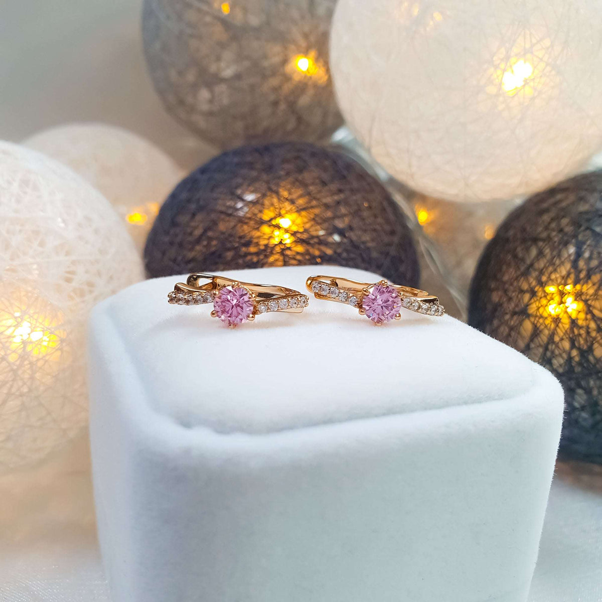 Pink | Pair of gold-plated pink cubic zirconia earrings on a white box, warm lights behind