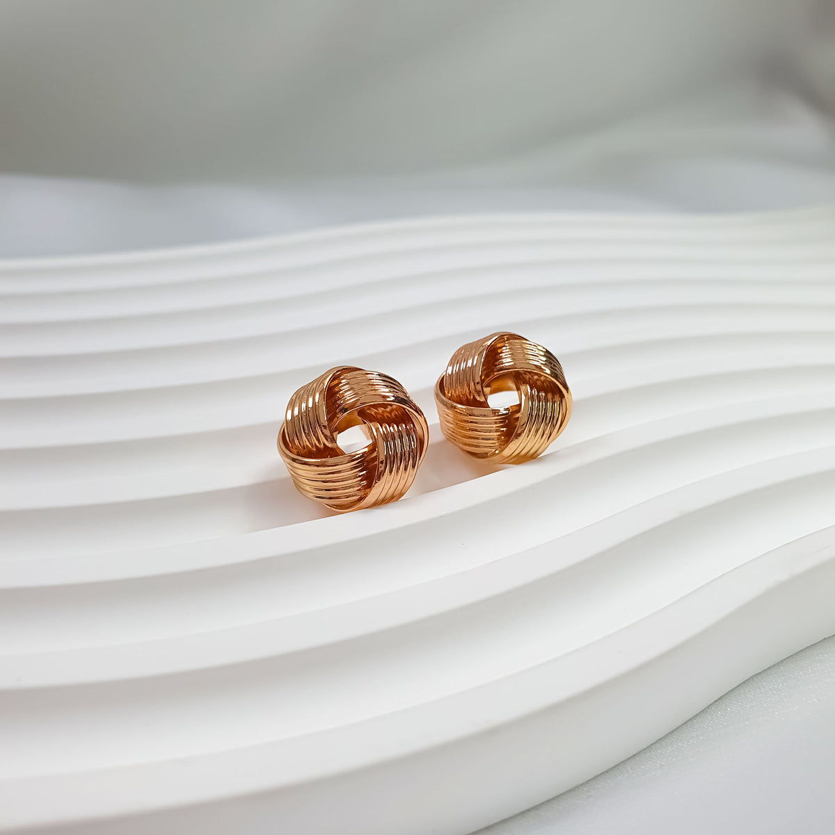 Knot stud earrings with textured grooves and warm gold plating on a wavy background