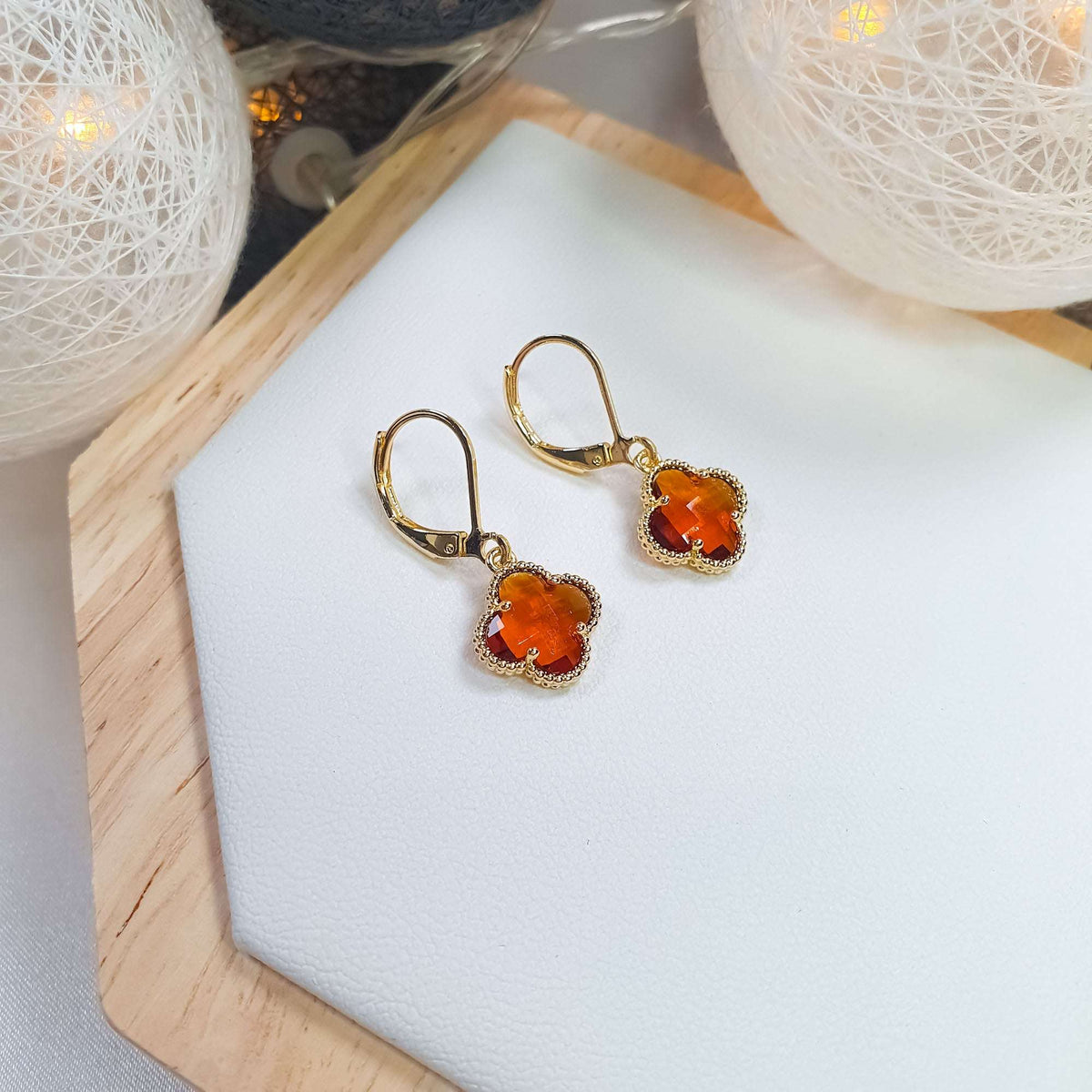 Pair of gold-plated earrings with orange four-leaf stones on a white stand, lights behind