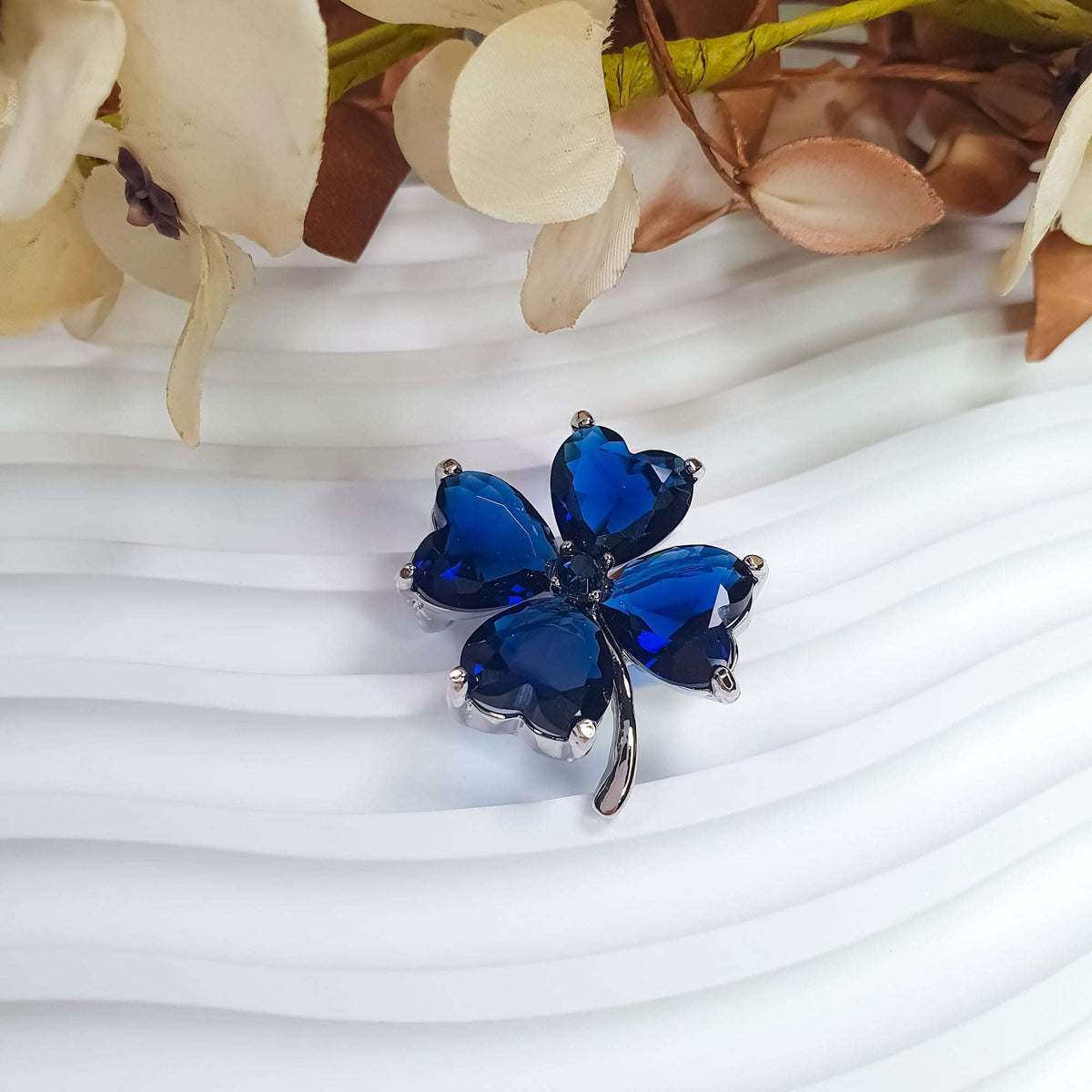 Dark blue clover brooch at an angle on a wavy background beside fabric flowers