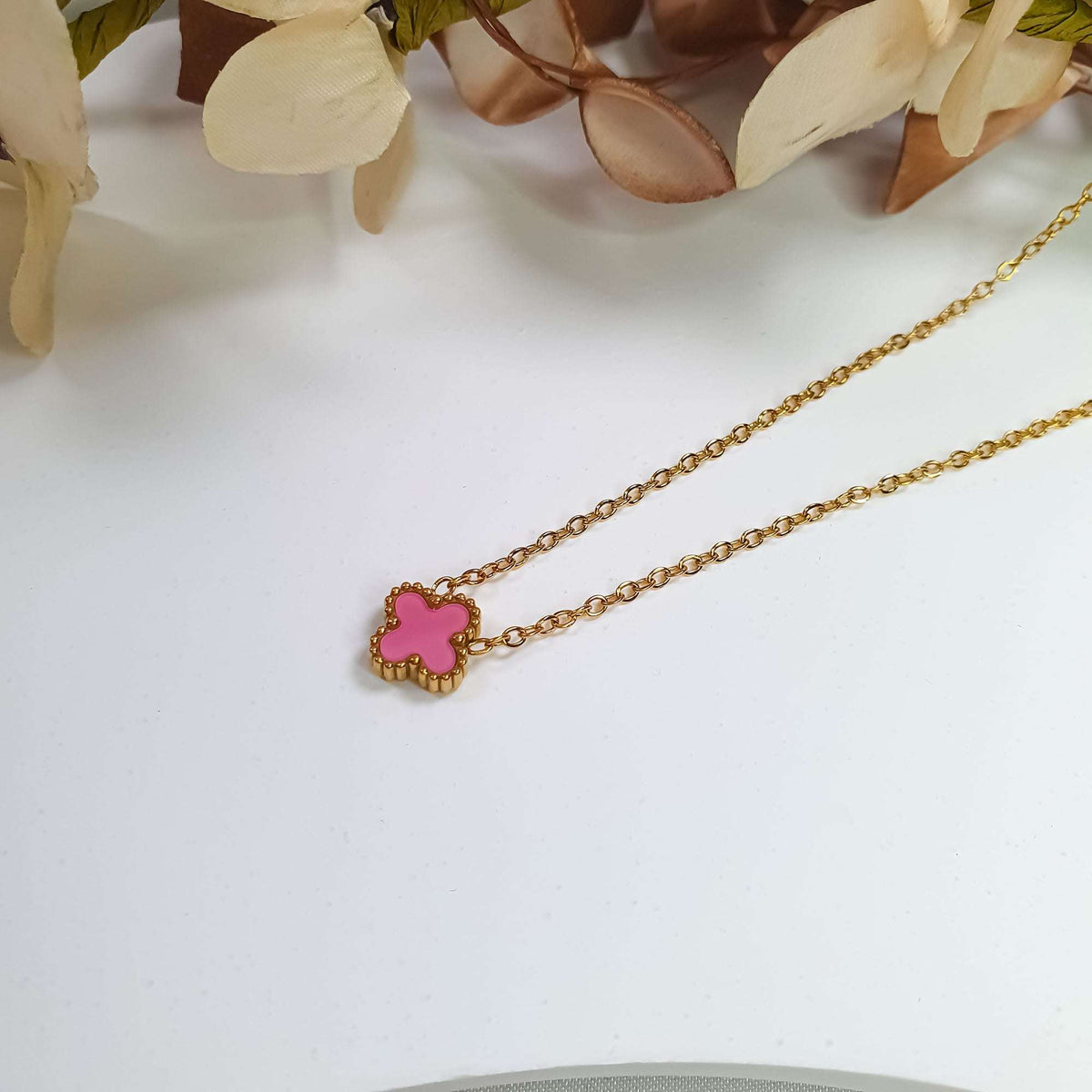 Pink enamel clover pendant close up on a gold-plated chain beside flowers