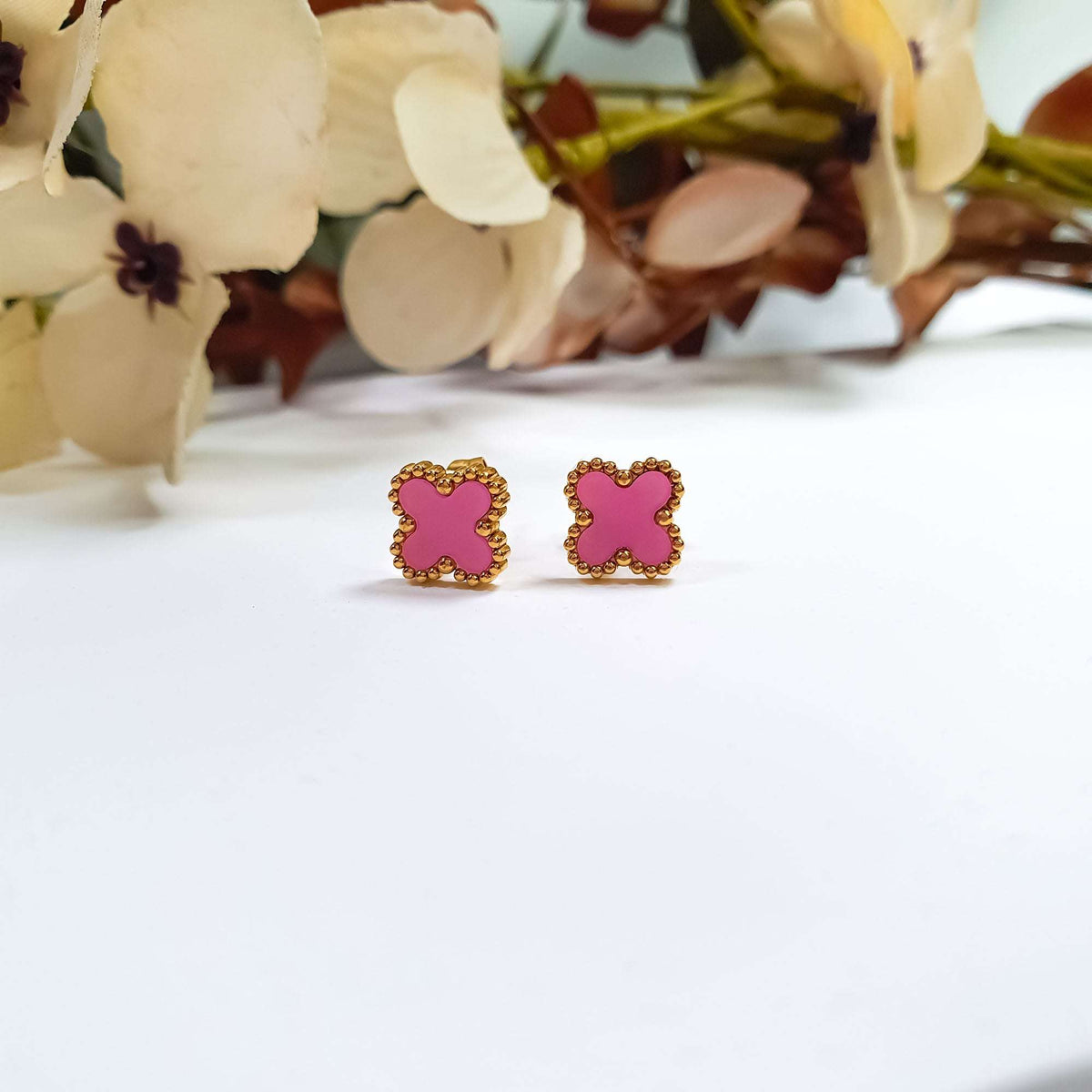 Small clover earrings with rich pink enamel and gold-tone beads, flowers behind