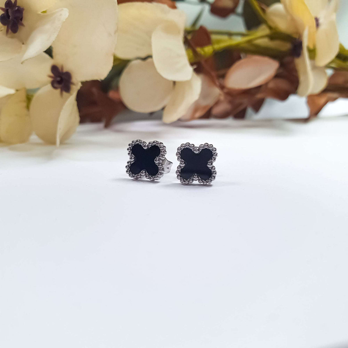 Pair of black clover studs with steel-tone beading beside dried flowers