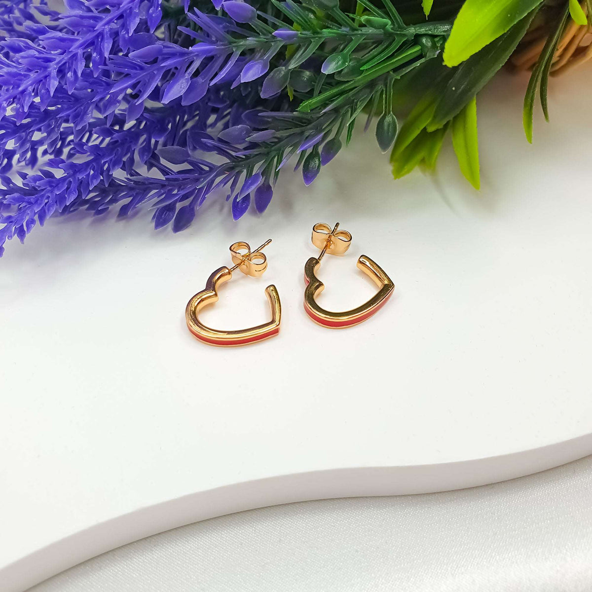 Pair of gold-plated heart outline earrings with red enamel beside lavender