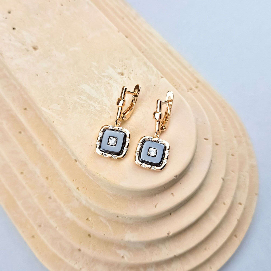Square gold-plated earrings with a ceramic inlay and central stone on a sand-coloured stand