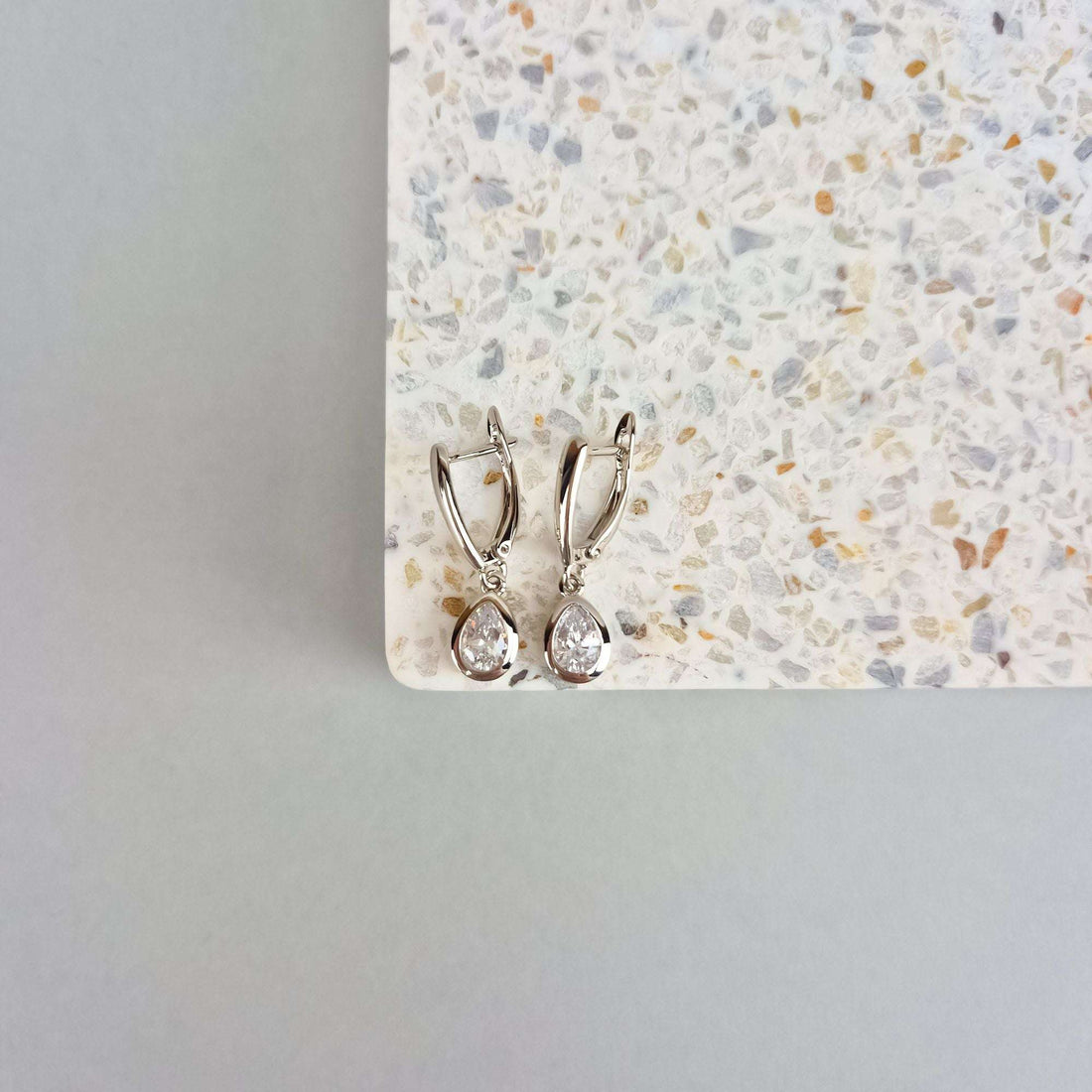 Silver-tone teardrop earrings on a pale speckled stone tile, close-up